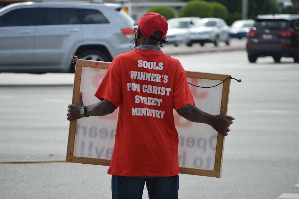 Street preacher in red shirt holding sign for Christian ministry.